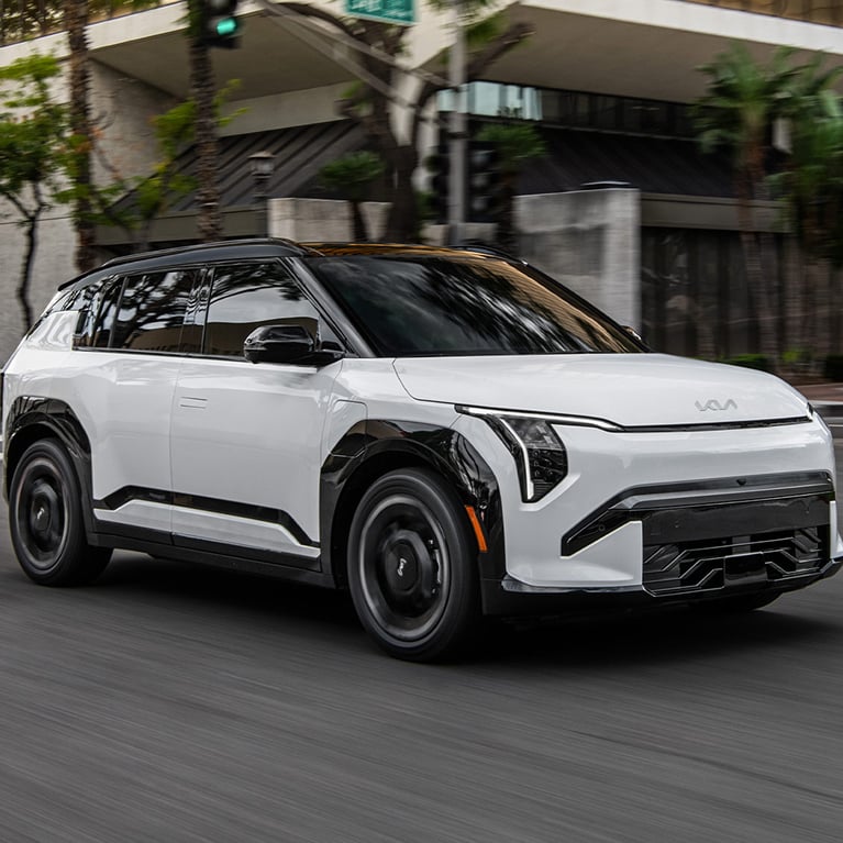 The 2027 Kia EV3 electric SUV GT-Line trim in white driving down an urban road with trees and interesting architecture.