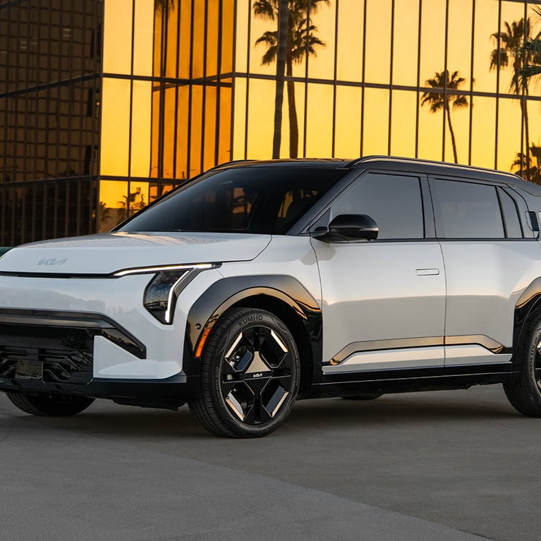 Front side angle photograph of the 2027 Kia EV3 electric compact SUV, showing silhouette, tires, headlights, door handles, and other exterior features of the electric vehicle. The EV3 is parked on asphalt with a golden glass skyscraper and palm trees in the background.