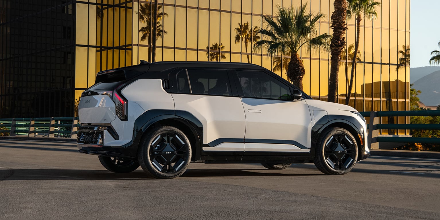 Exterior rear view of the 2027 Kia EV3 SUV parked on asphalt in an urban setting, with a glass building reflecting golden sunlight amplifying the striking silhouette of the SUV, including its rear trunk and black highlighting.
