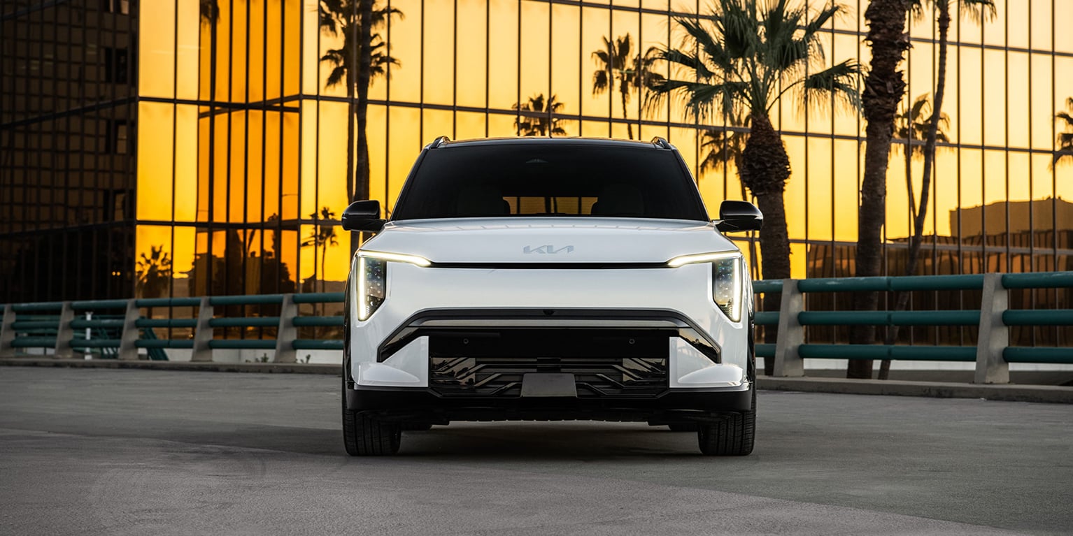 Straight-on front view of the 2027 Kia EV3 compact electric SUV, highlighting its headlights, grille, and overall shape. It is parked on asphalt with a golden skyscraper and palm trees behind it.