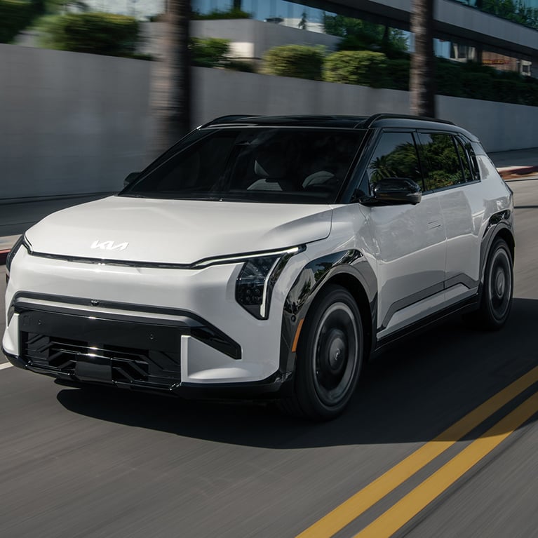 Dynamic shot of the 2027 Kia EV3 compact electric SUV in white driving rapidly down an urban road, with motion-blurred buildings and palm trees in the background. This photograph is meant to portray the excitement and fun of driving the EV3.