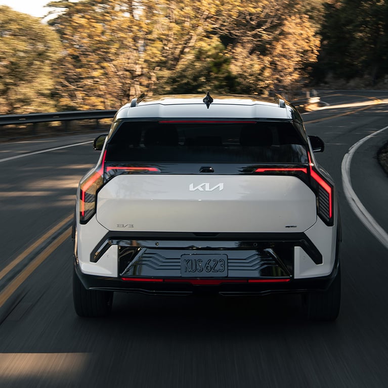 Straight-on photograph of the rear of the 2027 Kia EV3 compact electric SUV showcasing the back windshield, closed trunk door, and license plate. The EV is driving through a twisting road surrounded by rocks and trees, with sun and shadows across the asphalt.