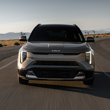 Front view of the 2027 Kia Niro Hybrid HEV sport utility vehicle as it drives around a curve on a desert road; the image highlights its LED projector headlights glowing white.