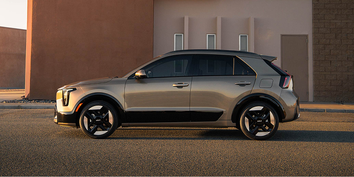 Side profile view of the 2027 Kia Niro Hybrid subcompact SUV in a bronze brown color, highlighting its distinctive shape and geometric tire designs. The EV is parked in front of a building in the desert.