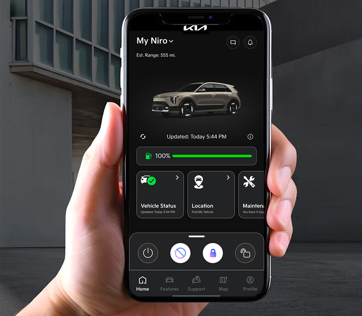 A hand holding a cellular phone, which has the Kia Connect app onscreen. The app is displaying a Kia vehicle and various status and location information. Building architecture is visible in the background.