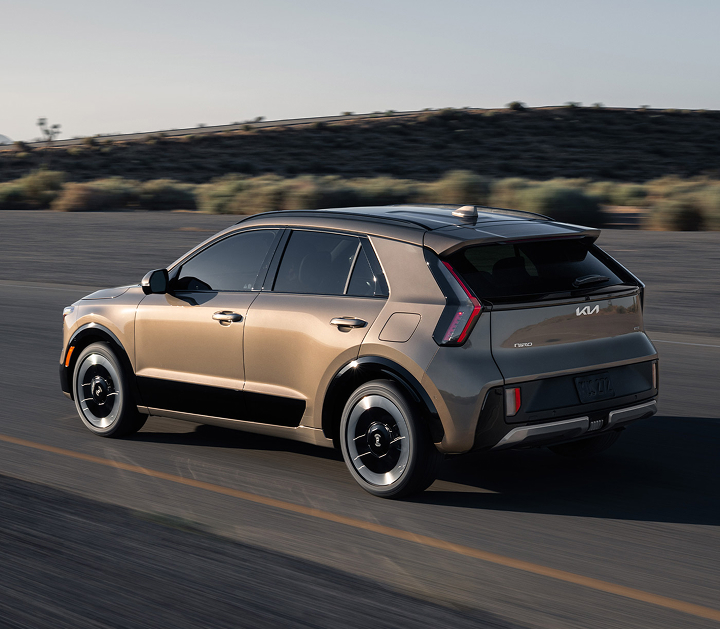 Rear angle photograph of the 2027 Kia Niro Hybrid Electric SUV driving over an outdoor road with desert vegetation in the background.