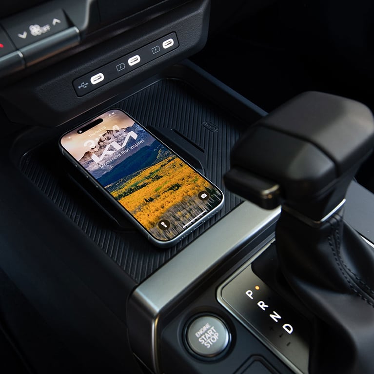 Close-up interior view of the 2027 Kia Seltos gear shifter in park and wireless charging station holding a charged smartphone with the Kia logo and mountains as the lock screen, highlighting the modern tech features.
