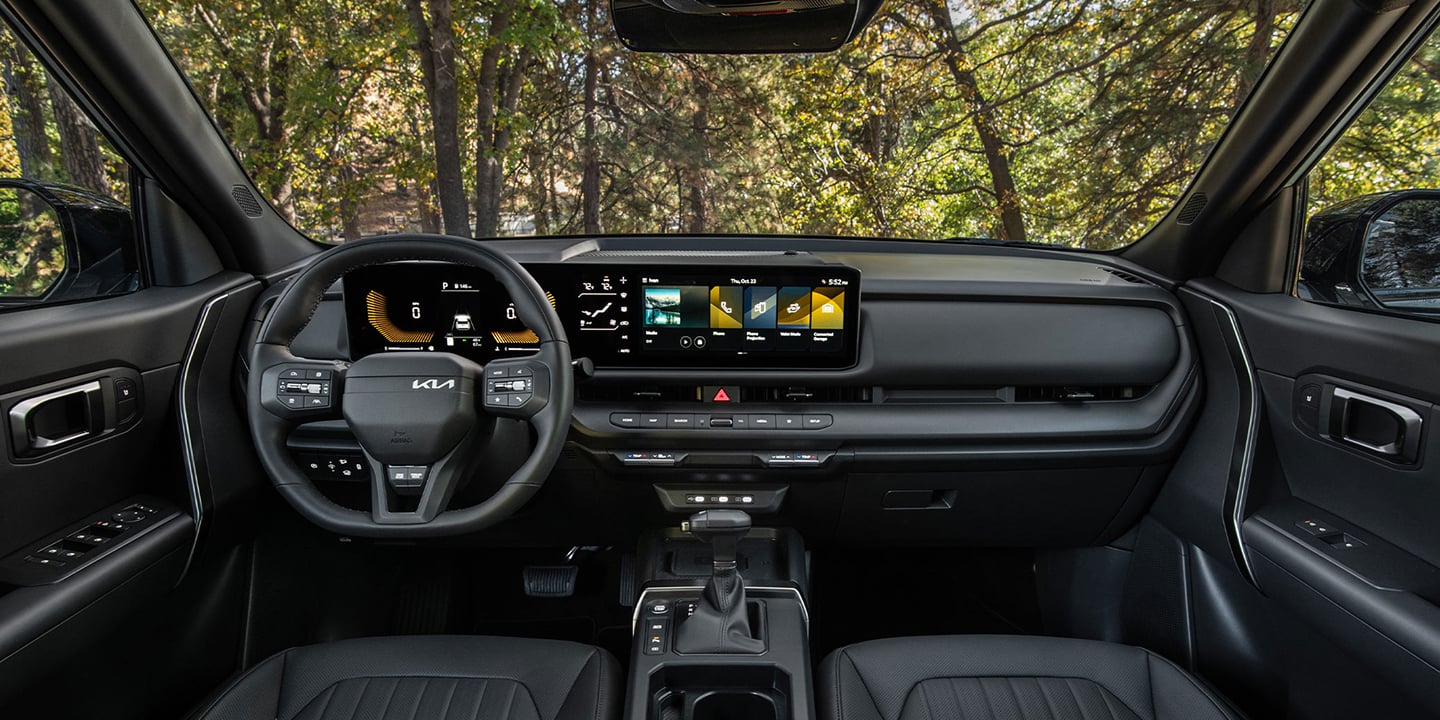 Interior wide-angle photo inside the 2027 Kia Seltos showcasing its steering wheel, dashboard display, center console, shifter, rear view mirrors, glove compartment, and more. A forest is visible through the windshield.
