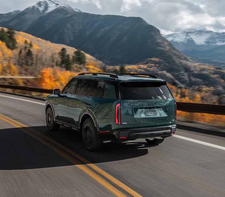 An action shot of the 2027 Kia Telluride Hybrid X-Line SX Prestige in blue-green driving on a highway, with a dense forest and snowy mountains in the background. The image highlights the rear and driver-side view of the SUV.
